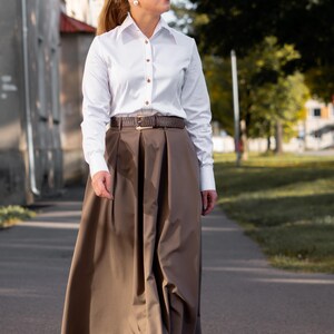 Flat lay of the white blouse, highlighting button and stitching details from a top-down view. This image also confirms that the shirt comes with brown designer buttons and decorative contrast stitching. Accessories pictured are for styling only and not included.