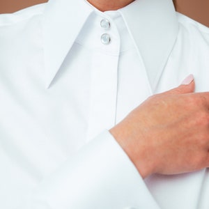 K&ouml;nnte beinhalten: Ein wei&szlig;es Langarmhemd mit einem hohen, strukturierten Kragen, der am Hals mit zwei runden, schimmernden Kn&ouml;pfen versehen ist. Die Manschette ist mit einem einzelnen, kunstvollen, silberfarbenen Zierknopf in Knotenoptik geschlossen.