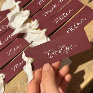 May include: A collection of rectangular burgundy paper place cards featuring white calligraphy names. Each card has a hole at the top with a white fabric ribbon tied through it. The names written on the cards include Dodge, Mark, Helen, Allan, Sarah, Keely, Christine, and Lee.