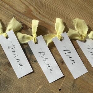 Hand holding a place card with elegant handwritten calligraphy in black ink. Butter Lemon Yellow Ribbon is tied though a gold eyelet in a linen frayed material

Wedding Place Cards, Baby Shower Place Cards, Handwritten Place Names, Luxury Wedding Place Cards, Lemon Yellow Ribbon, Butter Yellow Ribbon, Table Names, Pastel Yellow, Gender Neutral Place Cards