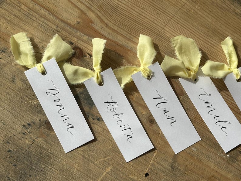 Hand holding a place card with elegant handwritten calligraphy in black ink. Butter Lemon Yellow Ribbon is tied though a gold eyelet in a linen frayed material

Wedding Place Cards, Baby Shower Place Cards, Handwritten Place Names, Luxury Wedding Place Cards, Lemon Yellow Ribbon, Butter Yellow Ribbon, Table Names, Pastel Yellow, Gender Neutral Place Cards