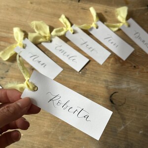Hand holding a place card with elegant handwritten calligraphy in black ink. Butter Lemon Yellow Ribbon is tied though a gold eyelet in a linen frayed material

Wedding Place Cards, Baby Shower Place Cards, Handwritten Place Names, Luxury Wedding Place Cards, Lemon Yellow Ribbon, Butter Yellow Ribbon, Table Names, Pastel Yellow, Gender Neutral Place Cards