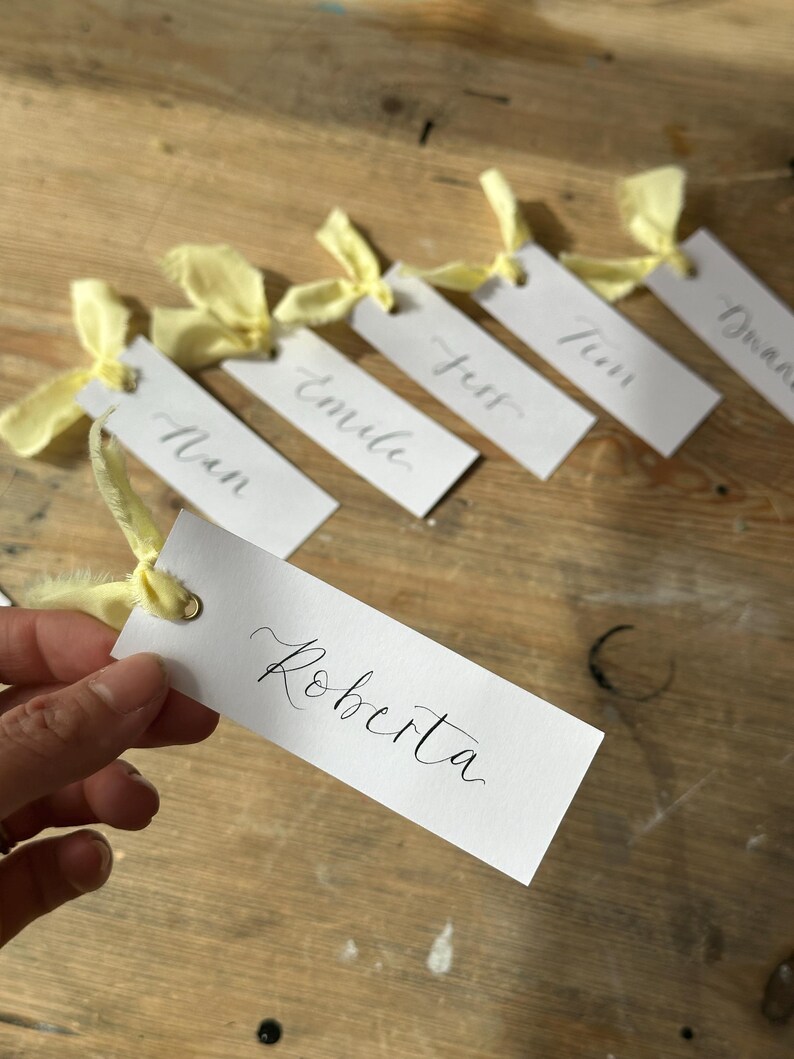 Hand holding a place card with elegant handwritten calligraphy in black ink. Butter Lemon Yellow Ribbon is tied though a gold eyelet in a linen frayed material

Wedding Place Cards, Baby Shower Place Cards, Handwritten Place Names, Luxury Wedding Place Cards, Lemon Yellow Ribbon, Butter Yellow Ribbon, Table Names, Pastel Yellow, Gender Neutral Place Cards