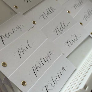 Kalligrafie DIY bruiloft plaatsnaamkaart | Witte kaart met handgeschreven kalligrafie, blanco platte plaatskaart gouden oogje lintgat