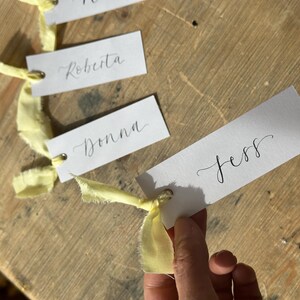 Hand holding a place card with elegant handwritten calligraphy in black ink. Butter Lemon Yellow Ribbon is tied though a gold eyelet in a linen frayed material

Wedding Place Cards, Baby Shower Place Cards, Handwritten Place Names, Luxury Wedding Place Cards, Lemon Yellow Ribbon, Butter Yellow Ribbon, Table Names, Pastel Yellow, Gender Neutral Place Cards
