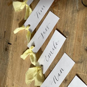 Hand holding a place card with elegant handwritten calligraphy in black ink. Butter Lemon Yellow Ribbon is tied though a gold eyelet in a linen frayed material

Wedding Place Cards, Baby Shower Place Cards, Handwritten Place Names, Luxury Wedding Place Cards, Lemon Yellow Ribbon, Butter Yellow Ribbon, Table Names, Pastel Yellow, Gender Neutral Place Cards