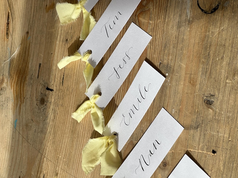 Hand holding a place card with elegant handwritten calligraphy in black ink. Butter Lemon Yellow Ribbon is tied though a gold eyelet in a linen frayed material

Wedding Place Cards, Baby Shower Place Cards, Handwritten Place Names, Luxury Wedding Place Cards, Lemon Yellow Ribbon, Butter Yellow Ribbon, Table Names, Pastel Yellow, Gender Neutral Place Cards