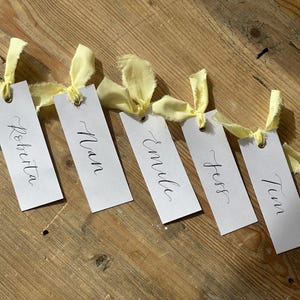 Hand holding a place card with elegant handwritten calligraphy in black ink. Butter Lemon Yellow Ribbon is tied though a gold eyelet in a linen frayed material

Wedding Place Cards, Baby Shower Place Cards, Handwritten Place Names, Luxury Wedding Place Cards, Lemon Yellow Ribbon, Butter Yellow Ribbon, Table Names, Pastel Yellow, Gender Neutral Place Cards