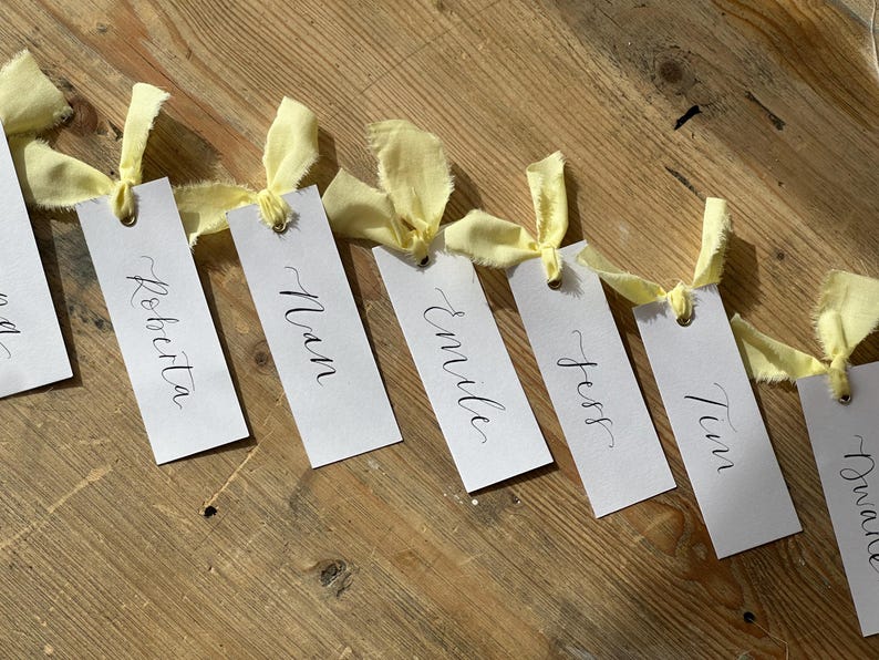 Hand holding a place card with elegant handwritten calligraphy in black ink. Butter Lemon Yellow Ribbon is tied though a gold eyelet in a linen frayed material

Wedding Place Cards, Baby Shower Place Cards, Handwritten Place Names, Luxury Wedding Place Cards, Lemon Yellow Ribbon, Butter Yellow Ribbon, Table Names, Pastel Yellow, Gender Neutral Place Cards