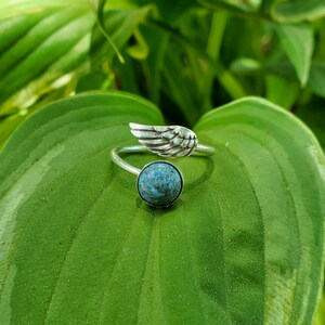 May include: A silver ring with a turquoise stone and a silver wing design. The ring is on a green leaf.