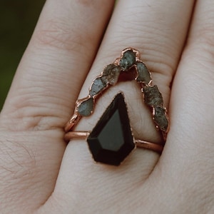 May include: A ring with a black gemstone set in a copper band. The band is adorned with a row of small, grey stones that form a V-shape.
