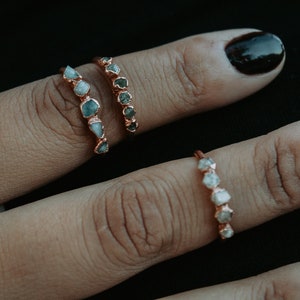 Op de afbeelding: Twee ringen met ruwe, ongeslepen edelstenen gezet in roségoud. De bovenste ring heeft vijf groene edelstenen, en de onderste ring heeft vijf heldere edelstenen.