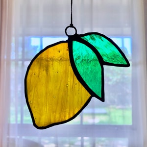 May include: A stained glass lemon ornament with a yellow lemon and green leaves. The ornament is hanging from a string.