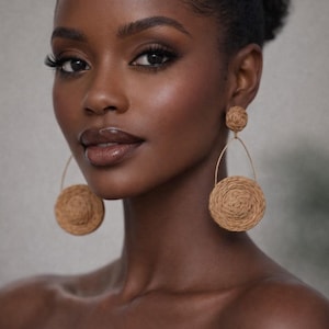 May include: A woman with dark skin and dark hair wears large, woven earrings. The earrings have a circular design, with a smaller circle at the top and a larger circle suspended below. The woman has dark eye makeup and glossy lips.