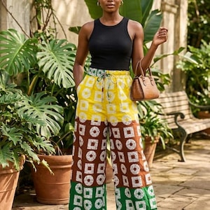 May include: A woman wearing a black tank top and patterned palazzo pants. The pants feature yellow, brown, and green geometric designs. She is holding a brown handbag and standing in a garden setting.