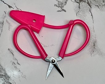 Könnte beinhalten: Rosa Nähschere mit einem kleinen Griff und einer Metallklinge. Die Schere hat ein einzigartiges Design mit einer Schlaufe oben zum einfachen Verstauen.