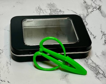 Könnte beinhalten: Eine schwarze Metalldose mit einem durchsichtigen Plastikfenster, die eine kleine, leuchtend grüne Schere enthält.