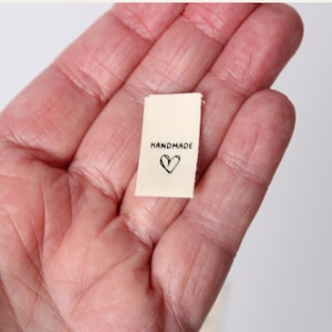 Pode incluir: Uma pequena etiqueta retangular de tecido em tom off-white com a palavra HANDMADE impressa em letras mai&uacute;sculas pretas acima de um contorno de cora&ccedil;&atilde;o preto desenhado &agrave; m&atilde;o. A etiqueta tem uma textura tecida e um design simples.