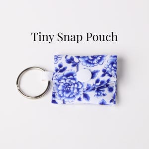 May include: A small, square snap pouch with a blue and white floral pattern, a white button closure, and a silver key ring. The text "Tiny Snap Pouch" is above the pouch.