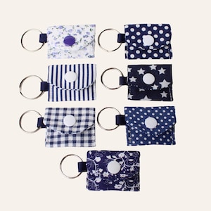 May include: A selection of fabric keyrings in diverse patterns, such as floral, polka dots, stripes, stars, and gingham. Each keyring has a small pouch with a snap closure and a metal ring. The colour scheme includes shades of blue, white, and purple.