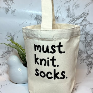 May include: A white canvas tote bag with black text that reads "must knit socks". The bag has a white fabric handle.