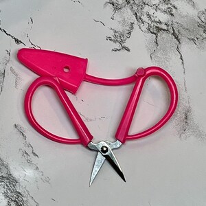 Puede incluir: Tijeras de coser de plástico rosa con una cuchilla plateada. Las tijeras tienen un protector de seguridad en la parte superior.