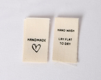 Könnte beinhalten: Zwei rechteckige, cremefarbene Stoffetiketten für Kleidung mit schwarzer Schrift. Das linke Etikett zeigt das Wort HANDMADE über einem handgezeichneten Herz. Das rechte Etikett trägt den Text HAND WASH und LAY FLAT TO DRY.