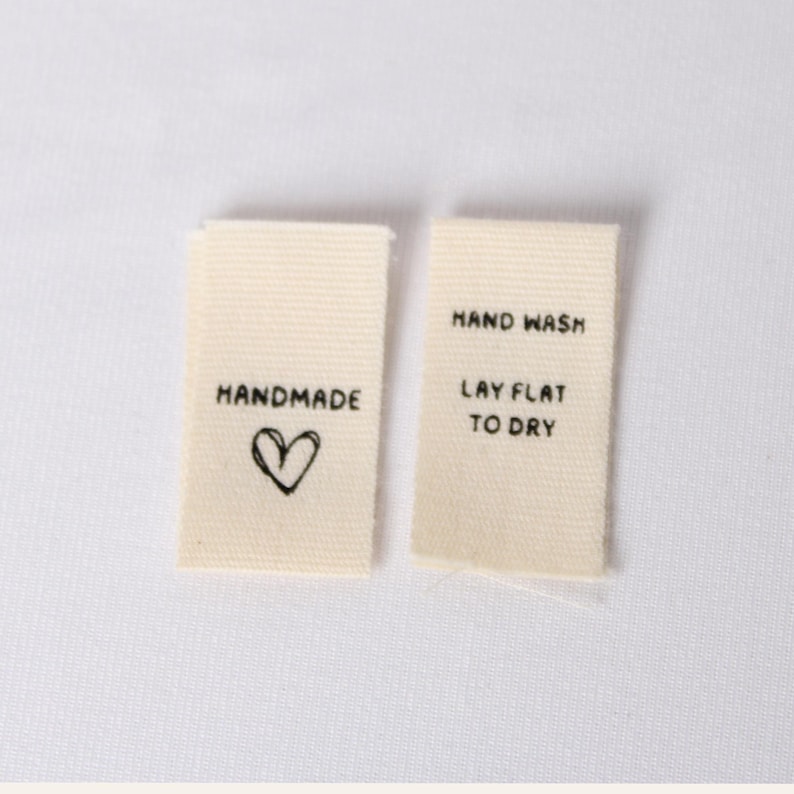 Pode incluir: Duas etiquetas retangulares de tecido em tom off-white para roupas com texto em preto. A etiqueta esquerda apresenta a palavra HANDMADE acima de um cora&ccedil;&atilde;o desenhado &agrave; m&atilde;o. A etiqueta direita exibe o texto HAND WASH e LAY FLAT TO DRY.