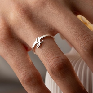 May include: A silver ring with the letter "g" in a cursive font. The ring is on a person's finger, with a white vase in the background. The ring is a delicate piece of jewelry.