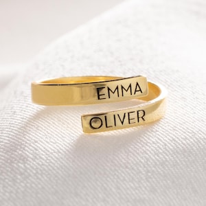 May include: A gold-coloured wrap ring engraved with the names "EMMA" and "OLIVER". The ring is presented on a white, textured surface. The design is modern and minimalist. The ring's simple design makes it a versatile piece of jewellery.