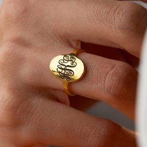 May include: A gold-colored signet ring with a black monogram on an oval face. The ring is worn on a finger, and the background is blurred, suggesting a focus on the jewelry.