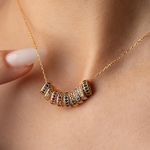 May include: A gold necklace featuring a series of colorful, gem-studded rings. The rings are arranged in a curved line, with each ring showcasing a different color of gemstones. The necklace is worn around the neck.