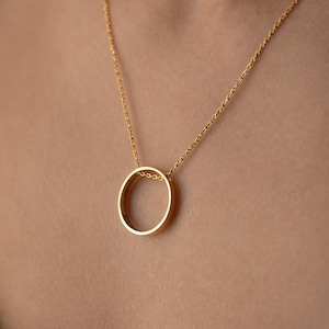 Könnte beinhalten: Eine goldfarbene Halskette mit feiner Kette und einem kreisförmigen Anhänger. Der Anhänger ist ein einfacher, offener Kreis. Die Halskette wird vor einem dunklen Hintergrund getragen.