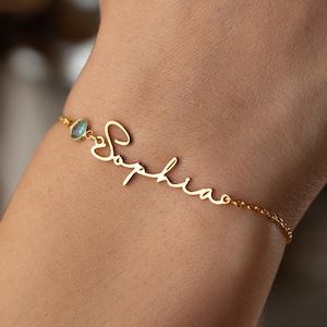 May include: A gold-colored bracelet with the name "Sophia" in cursive script. The bracelet features a small, light blue gemstone and a delicate chain. The bracelet is worn on a wrist.