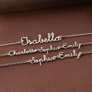 May include: Three silver name necklaces. The top necklace spells "Isabella" in cursive script. The second necklace spells "Charlotte Sophia Emily". The third necklace spells "Sophia Emily". The necklaces are on a brown surface.