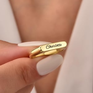 Peut inclure: Une bague dorée polie avec une surface supérieure plate et rectangulaire. Le prénom Charlotte est gravé en écriture cursive noire au centre de la surface plate. L'anneau est lisse, arrondi et présente une finition métallique réfléchissante.