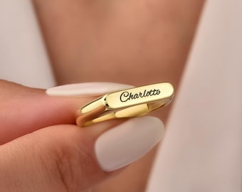 May include: A polished gold-tone ring featuring a flat, rectangular top surface. The name Charlotte is engraved in a black, cursive script font across the center of the flat surface. The band is smooth, rounded, and has a reflective metallic finish.