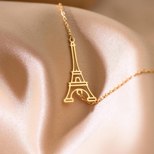 Könnte beinhalten: Eine goldfarbene Halskette mit einem Anhänger in Form des Eiffelturms. Der Anhänger ist eine detaillierte Umrisszeichnung des berühmten Wahrzeichens. Die Halskette wird auf einem weichen, pfirsichfarbenen Stoff präsentiert.