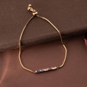May include: Gold-toned bracelet with a delicate chain and a row of colorful rectangular stones. The stones include blue, purple, red, and white. The bracelet is displayed on a brown surface.