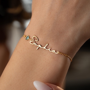 May include: A delicate gold bracelet with the name "Sophia" in cursive script. The bracelet features a small, light blue gemstone and a fine chain. The bracelet is designed to be worn on the wrist.