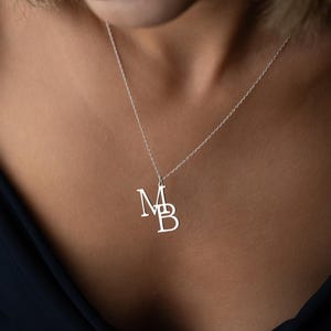 Peut inclure: Un collier argenté avec un pendentif représentant les lettres entrelacées "MB". Le pendentif est suspendu à une fine chaîne, posé sur la peau d'une personne. Le collier est un bijou simple et élégant.