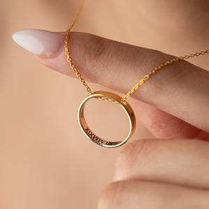 Könnte beinhalten: Eine goldfarbene Halskette mit einem kreisförmigen Anhänger. Der Anhänger hat eingravierte römische Ziffern und hängt an einer zarten Kette. Die Halskette wird von einer Hand gehalten, mit einem unscharfen Hintergrund.