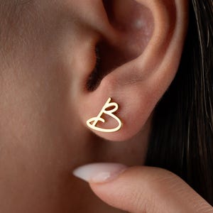 May include: A close-up of a gold-colored earring in the shape of the letter "B" is shown on an ear. The earring is a minimalist design, with a continuous line forming the letter. The skin tone is light.