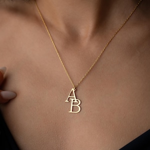 May include: A gold-colored necklace with the letters "AB" as a pendant. The pendant is suspended from a delicate chain. The necklace is displayed against a dark background.