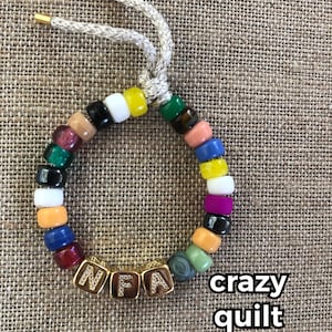 May include: A colorful beaded bracelet with gold letter beads spelling "NFA". The bracelet is made with a variety of colors, including red, orange, yellow, green, blue, and purple. The bracelet is tied with a beige cord.
