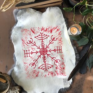 May include: A white, irregularly shaped paper with a red, stylized symbol at the center. The symbol is surrounded by a red, textured border. The paper rests on a gray, furry surface, with a skull and other objects in the background.
