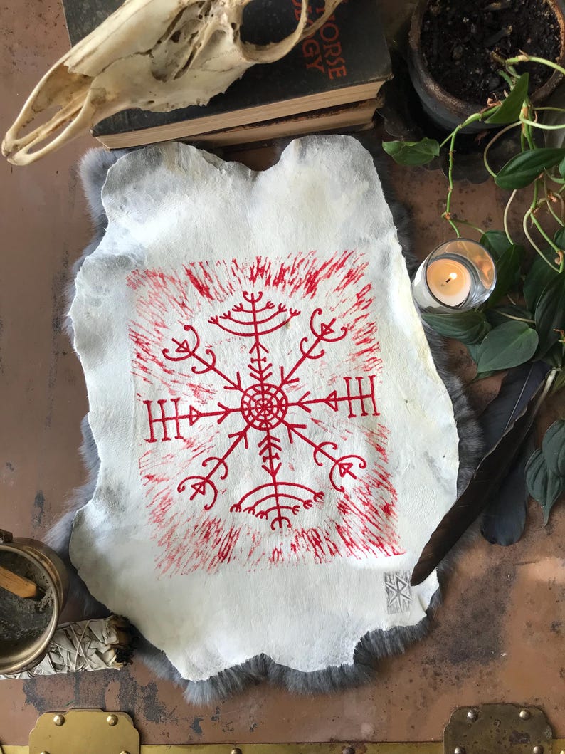 May include: A white, irregularly shaped paper with a red, stylized symbol at the center. The symbol is surrounded by a red, textured border. The paper rests on a gray, furry surface, with a skull and other objects in the background.