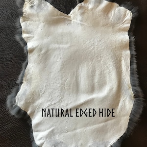 May include: A natural-edged hide with a white interior and a gray fur border. The hide is irregularly shaped and has the text "NATURAL EDGED HIDE" printed on it. The hide is displayed on a dark brown surface.