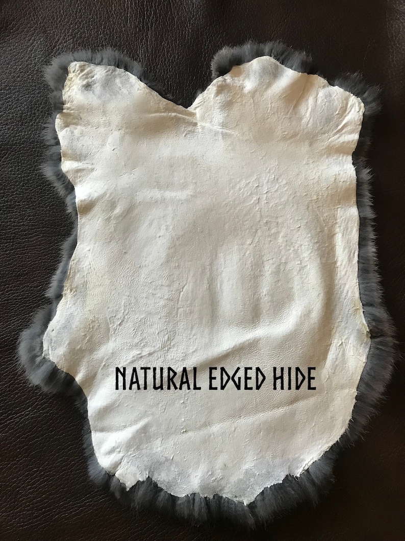 May include: A natural-edged hide with a white interior and a gray fur border. The hide is irregularly shaped and has the text "NATURAL EDGED HIDE" printed on it. The hide is displayed on a dark brown surface.