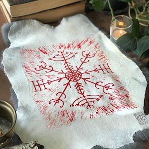 May include: A white, textured hide with a red, stylized snowflake design. The artwork features a central circular element with radiating lines and symbols. The hide is displayed on a gray, furry surface, with other items in the background.